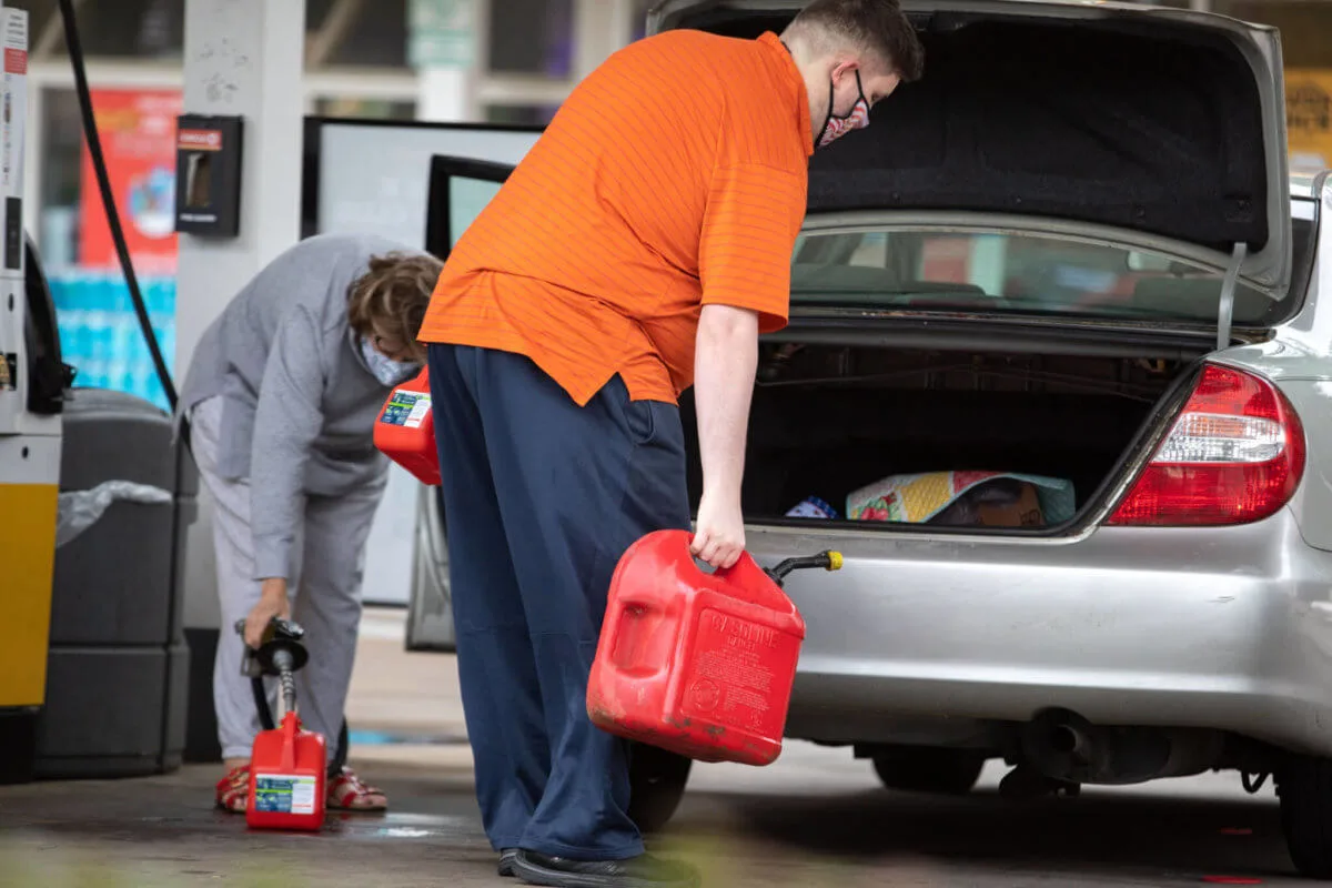 A person in an orange shirt accompanied by another in gray load numerous gas containers into a sedan at a Charlotte, NC gas station.