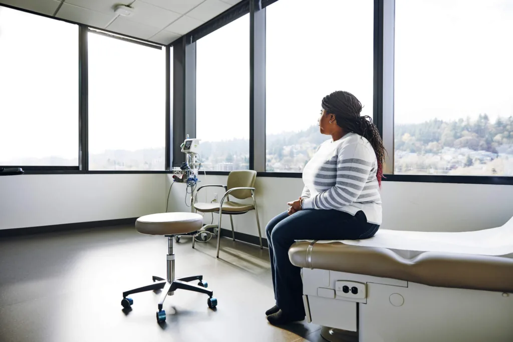 A dark-haired woman sits on the examination table in a doctor's office, looking away to the left.