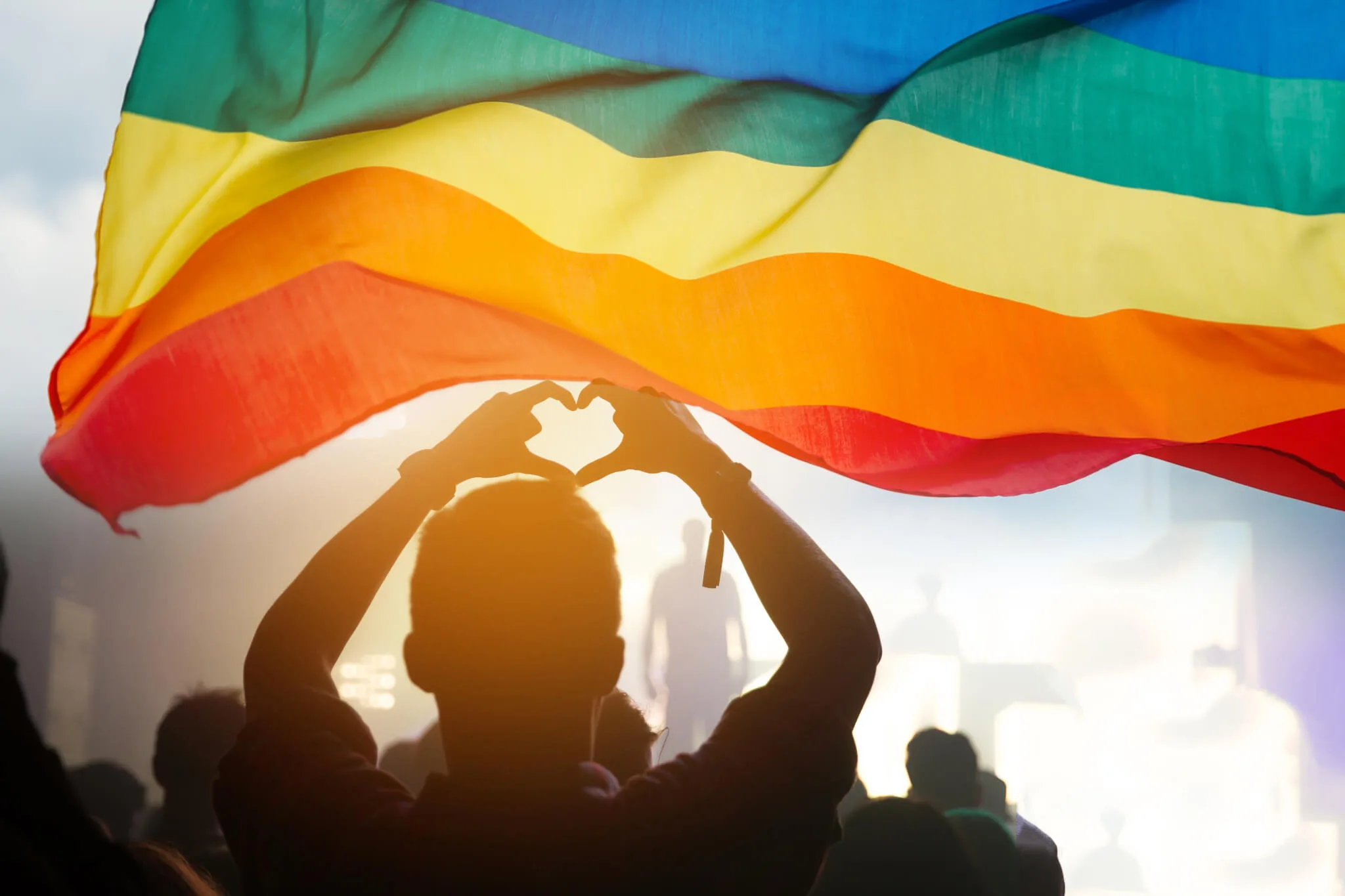 A person backlit by the sun makes a shape of a heart with their fingers while standing beneath a rainbow flag.