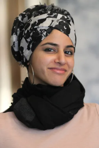 an olive-complexioned person wears a black and white scarf on their hair, hoop earrings, and a black scarf around their neck. They are looking fdirectly at the xas
