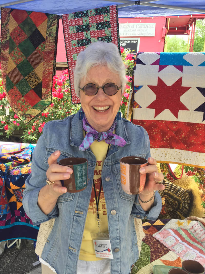 Grey[haired person in sunglasses and denim jacket stands before a quilt display holding up two mugs in welcome.