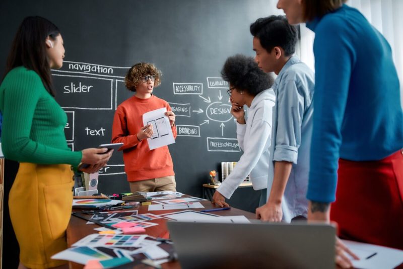 Young professional designer is showing some sketches with mobile app web design to his colleagues while having a meeting in the modern office. Working together. Teamwork.