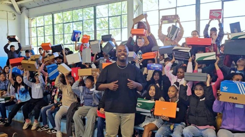 Photo of a teen posing in front of several of children holding up brand new shoe boxes.