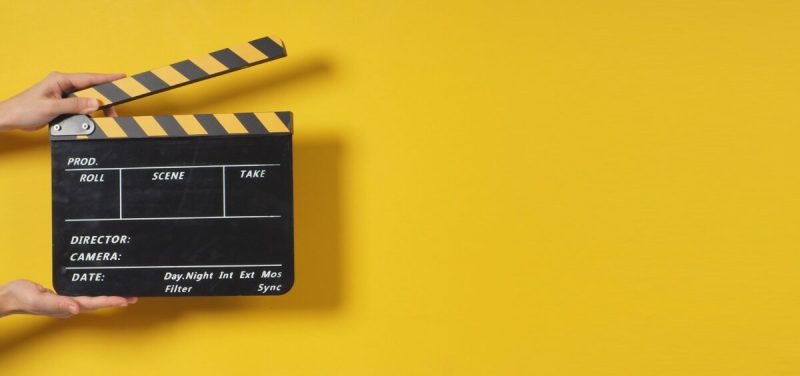 a yellow background with a hand holding a black clapper board used for movies.