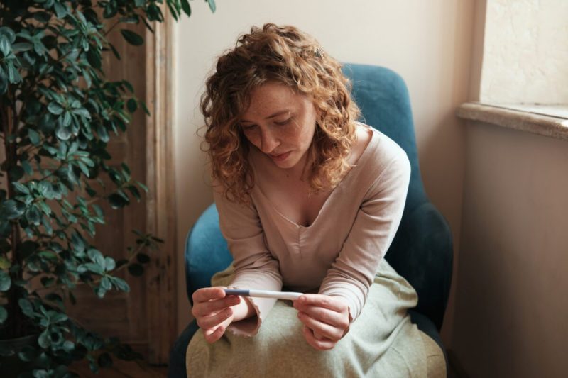 Photo of a woman sitting on a chair looking worried while holding a positive pregnancy test.