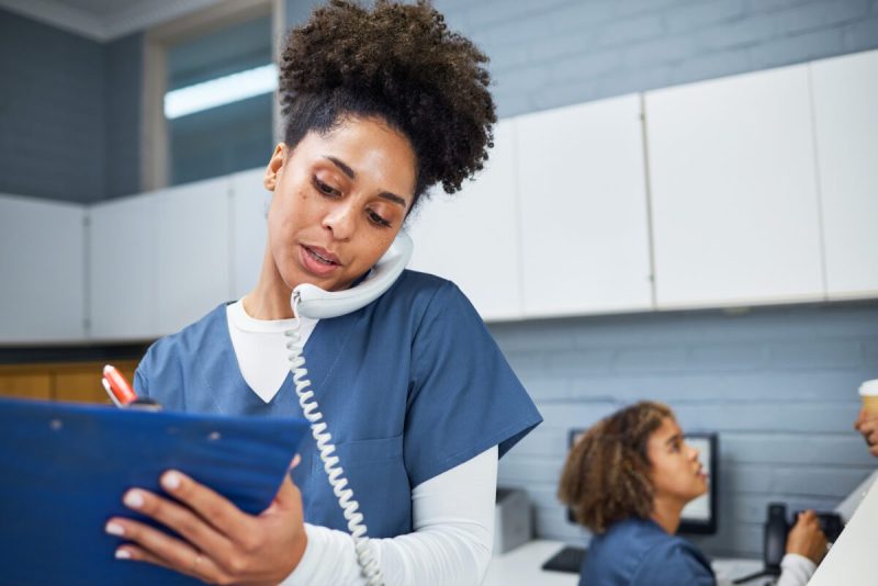 Woman nurse writes on a clipboard while answering the phone at a front desk. A nurse behind her, sits down in front of a computer at a table.