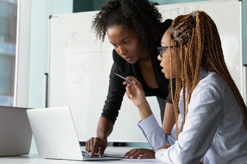 Photo of two woman working. One woman is leaning over to help the other woman, sitting down, on the computer.