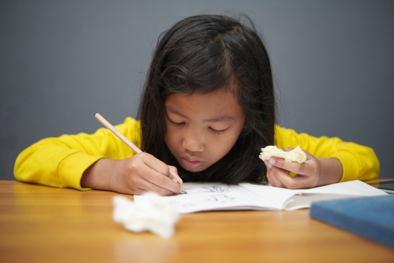 Photo of a little girl doing schoolwork while holding a half-eaten sandwich